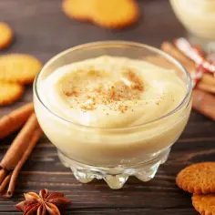 Kremowy pudding w szklanej miseczce posypany cynamonem, obok ciasteczka i laski cynamonu.