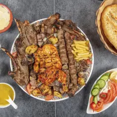 Talerz grillowanej baraniny, kurczaka, frytek, sałatki i sosów, obok chleba.